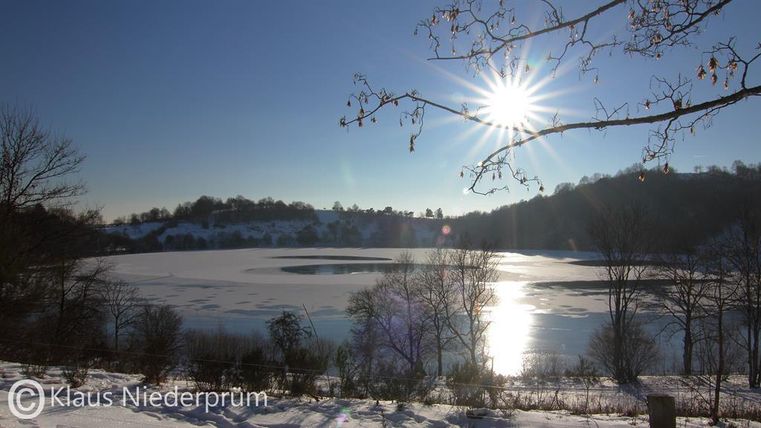 Een winterlandschap met een bevroren meer en met sneeuw bedekte oevers. De zon straalt helder aan de lucht en weerkaatst in het water.