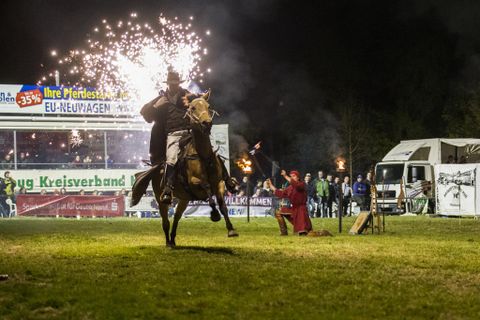 Ein Reiter auf einem Pferd zeigt eine spektakuläre Darbietung mit Feuerwerk im Hintergrund. Zuschauer beobachten das Ereignis in der Nacht.