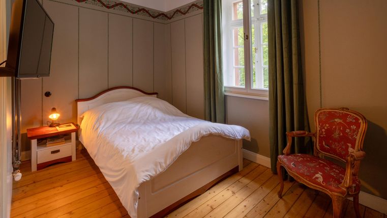 A cozy bedroom with a double bed and a television. Natural light filters through the window, and a red chair complements the decor.
