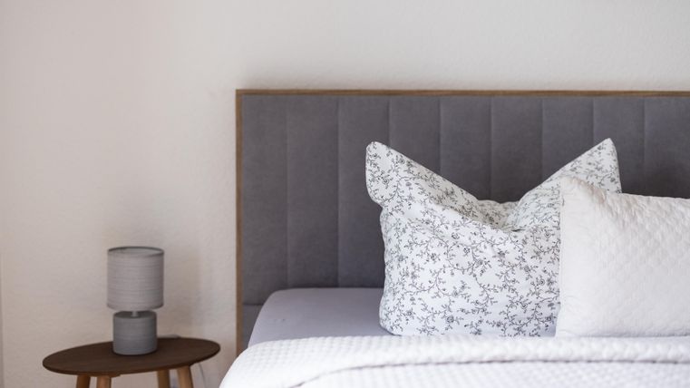 A modern bedroom with a gray headboard and elegant pillows. Next to the bed is a small table with a lamp.
