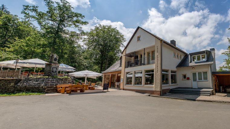 Ein modernes Restaurant mit einem schönen Außenbereich und Sonnenschirmen. Umgeben von grünen Bäumen und klarem Himmel.