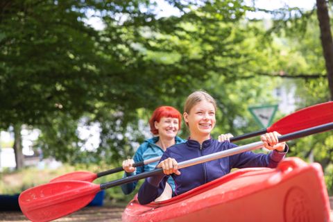 Zwei Personen sitzen in einem roten Kajak und paddeln fröhlich. Im Hintergrund sind Bäume und eine ruhige Umgebung zu sehen.