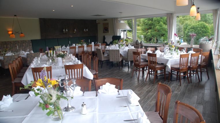 A bright restaurant with elegant wooden tables and chairs. The tables are neatly set, and there are windows overlooking the green surroundings.
