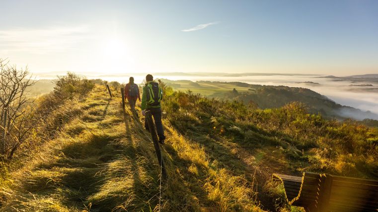 Zwei Wanderer gehen einen Pfad entlang einer hügeligen Landschaft. Im Hintergrund ist ein klarer Himmel und eine Nebelbank zu sehen.