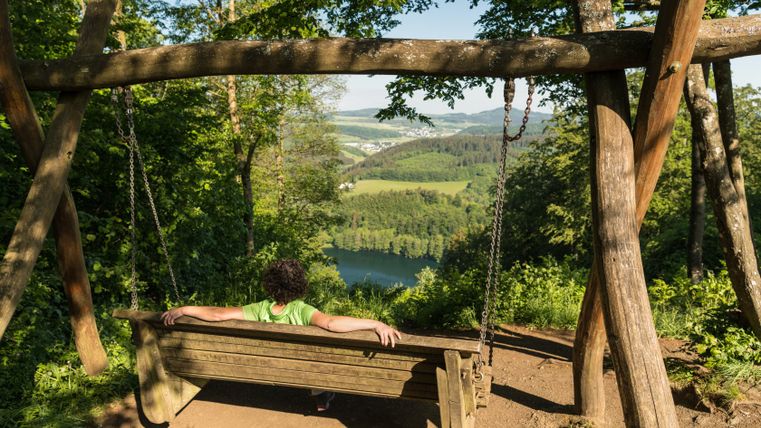 Person sitzt auf einer Holzschaukel mit Blick auf das Gemündener Maar, umgeben von grünen Bäumen und Landschaft.