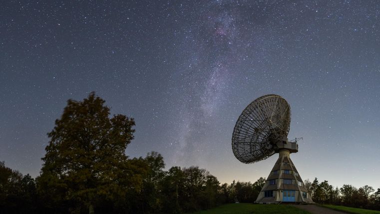 Ein großes Radioteleskop steht unter einem klaren Nachthimmel mit der Milchstraße. Bäume umgeben die Anlage in einer ruhigen Landschaft.