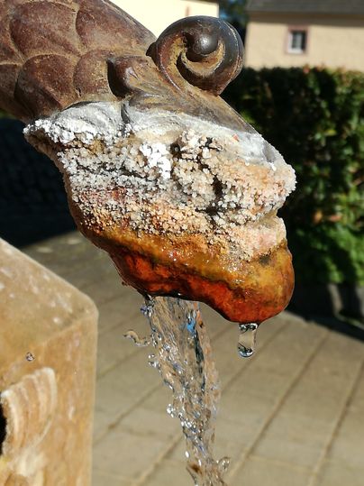 Ein Wasserhahn in Form eines Kopfes mit Mineralablagerungen. Wasser fließt aus dem Hahn und sprudelt herunter.