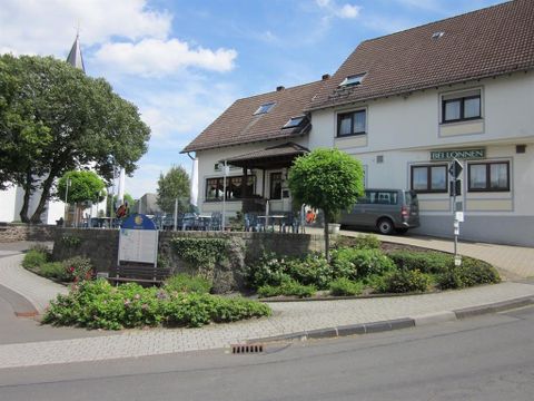 View from the front of the inn Bei Lonnen.
The two-story building is brightly painted and has a black tiled roof.
In front of the guesthouse, you can see a round terrace equipped with several tables and chairs.