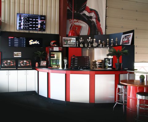 Ein modernes Verkaufsstand mit roten und schwarzen Elementen. Im Hintergrund sind Monitore und Pokale sichtbar, die auf eine Kartbahn hinweisen.