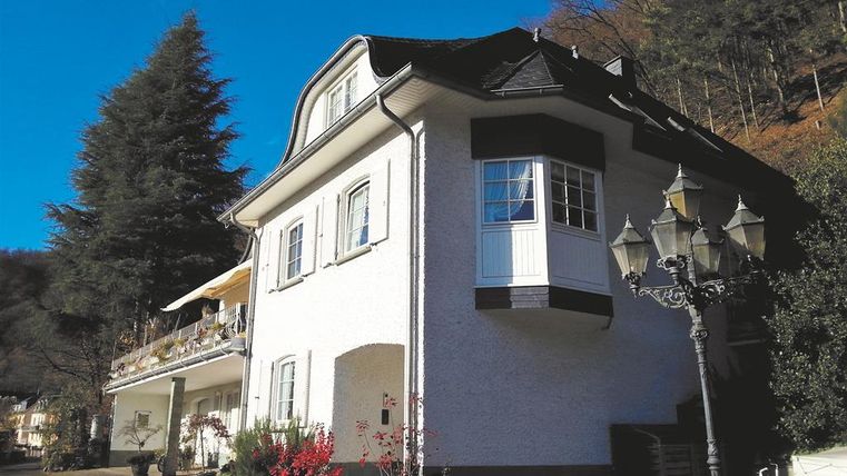 Ein charmantes Haus mit zwei Etagen und einem Balkon, umgeben von Bäumen und Blumen. Die Fassade ist weiß, und eine alte Laterne steht vor dem Gebäude.