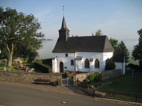 In the middle of the picture, you can see the small whitewashed chapel. The roof is covered with black slate. On the left side, there is a church tower with a pointed roof, also covered in black.
At the top of the church tower, you can see a weather vane. The chapel is surrounded by a churchyard that is framed by a rubble stone wall.