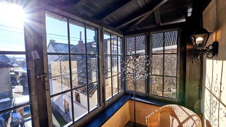 A cozy balcony with large windows and a seating area made of rattan. The sun shines through the windows, creating an inviting atmosphere.