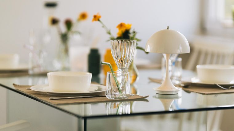 Een elegante eettafel met een glas en een moderne lamp. Op de tafel staan bloemen en servies in een lichte, uitnodigende sfeer.