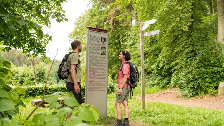 Two hikers stand in front of an information sign in the Weisshauswald forest, surrounded by green nature.