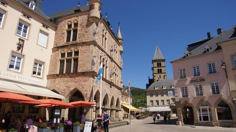 Ein malerischer Platz mit historischen Gebäuden und einem charmanten Café. Die Sonne scheint, und Menschen genießen die Atmosphäre.
