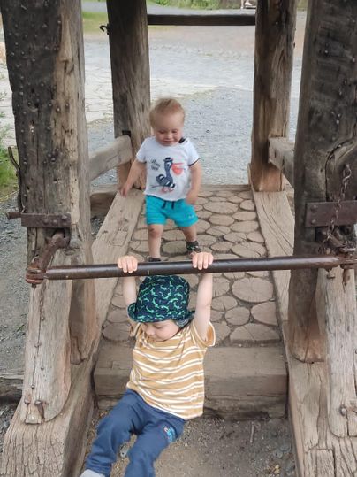 Two small children are playing on a playground. One is hanging from a wooden beam while the other stands next to it.
