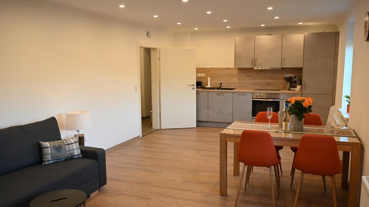 Een gezellig appartement met een moderne woonkamer en een open keuken. De eetruimte is voorzien van een houten tafel en oranje stoelen.