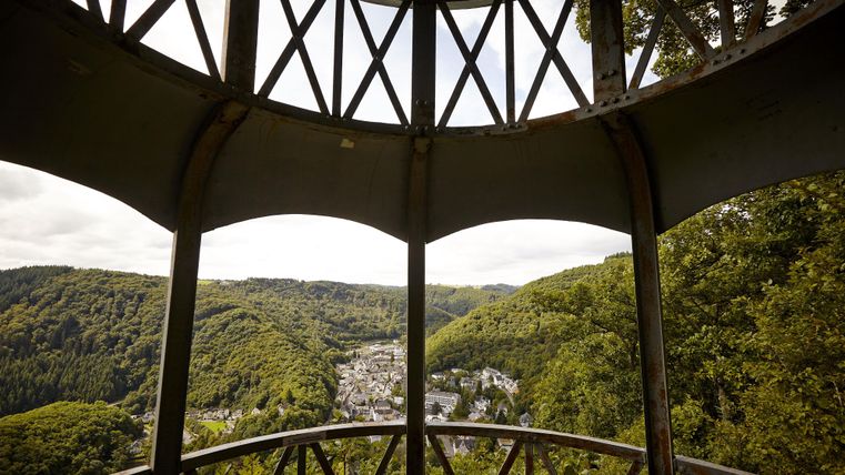 Ein Aussichtspunkt mit einem Gitterdesign, das einen Blick auf grüne Hügel und ein kleines Dorf im Tal bietet. Der Himmel ist bewölkt, was eine ruhige Atmosphäre schafft.
