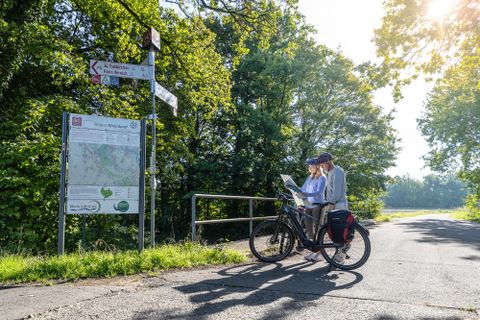 Twee fietsers staan bij een wegwijzer in het groen en bestuderen een kaart. De zonnige dag en de dichte bomen creëren een ontspannen sfeer.