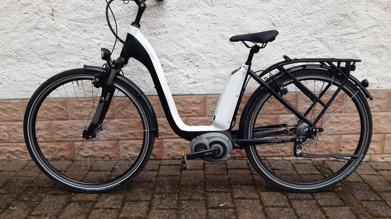 Ein modernes E-Bike in Schwarz und Weiß, mit niedrigem Einstieg und Gepäckträger. Es steht auf einem gepflasterten Untergrund vor einer hellen Wand.
