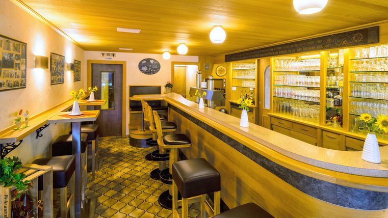 Eine einladende Bar mit Holzinterieur und beleuchteten Regalen. Die Sitzgelegenheiten sind komfortabel und es stehen frische Blumen auf den Tischen.