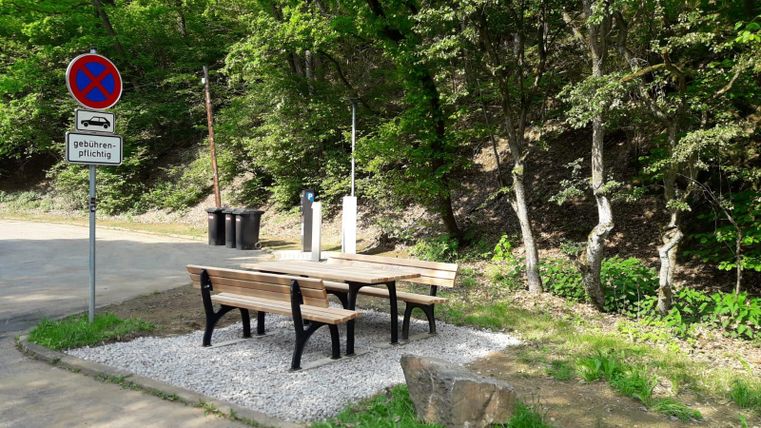 Ein Tisch mit Bänken steht in einer ruhigen, grünen Umgebung. In der Nähe gibt es ein Verkehrszeichen und Mülltonnen.