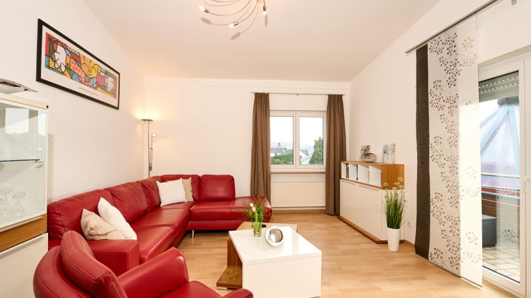 Ein modernes Wohnzimmer mit rotem Sofa und einem weißen Couchtisch. Große Fenster sorgen für viel Tageslicht und eine freundliche Atmosphäre.