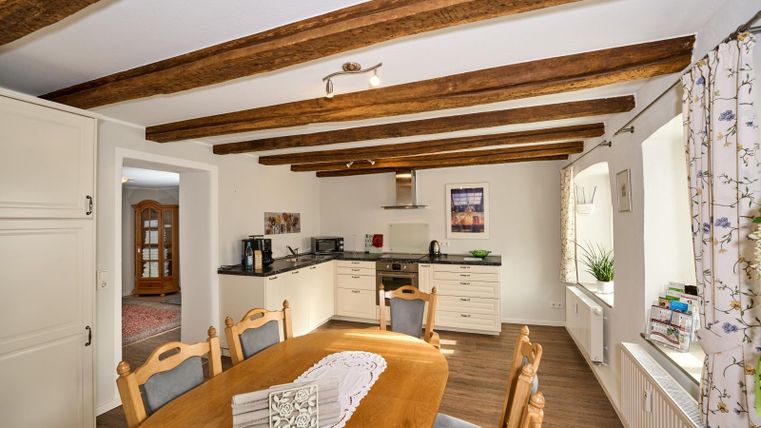 Een lichte keuken met houten balkenplafond en een grote eettafel. De moderne inrichting en ramen met uitzicht naar buiten creëren een uitnodigende sfeer.