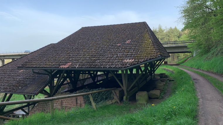 Ein schief stehendes Dach eines alten Gebäudes, umgeben von grünen Wiesen. Im Hintergrund verläuft ein Weg und eine Brücke.