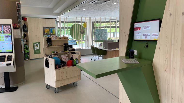 Ein moderner Empfangsbereich mit einem grünen Tisch und interaktiven Bildschirm. Die Wände sind hell und aus Holz, mit einer freundlichen Atmosphäre und verschiedenen Möbeln.