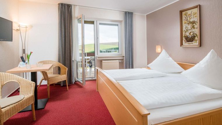 A cozy hotel room with two single beds. There is a table, two chairs, and a balcony with a view of the landscape.