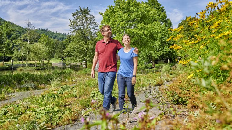 Ein Paar spaziert durch eine grüne Landschaft mit bunten Blumen. Im Hintergrund sind Bäume und ein ruhiger Teich zu sehen.