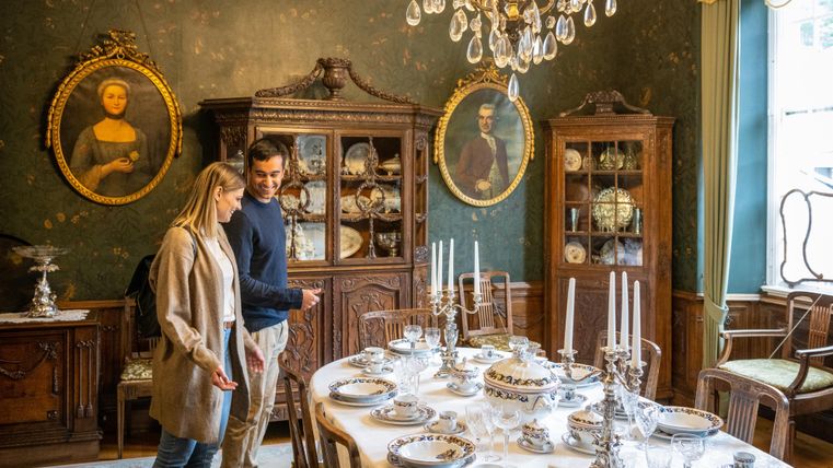 Een stijlvol ingerichte eetkamer met een elegante tafel, gedekt met fijn servies. Op de achtergrond zijn antieke meubels en portretten aan de wand te zien.