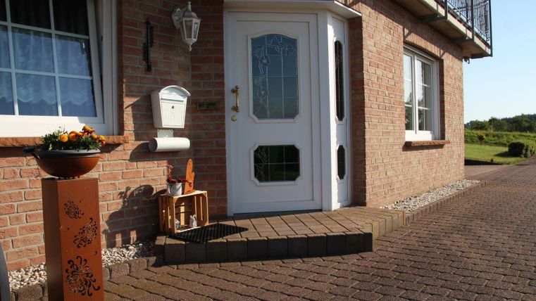 Ein einladender Eingangsbereich eines Hauses mit einer weißen Tür und einem kleinen Blumenständer. Die Fassade ist aus roten Ziegeln und es gibt einen gepflasterten Weg.