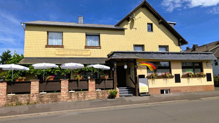 Ein gelbes Gebäude mit einem einladenden Restaurant und einer Terrasse. Parasols und eine deutsche Flagge sind sichtbar.