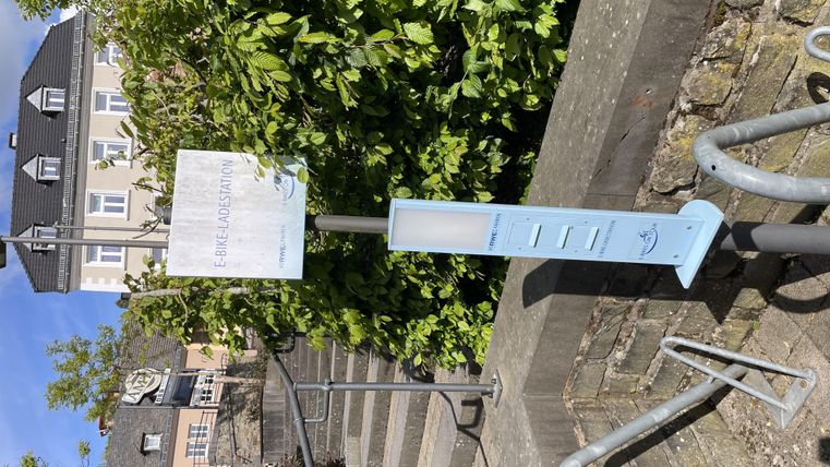 Een informatiezuil staat bij een trap, omringd door groene planten. Op de achtergrond zijn enkele huizen te zien.