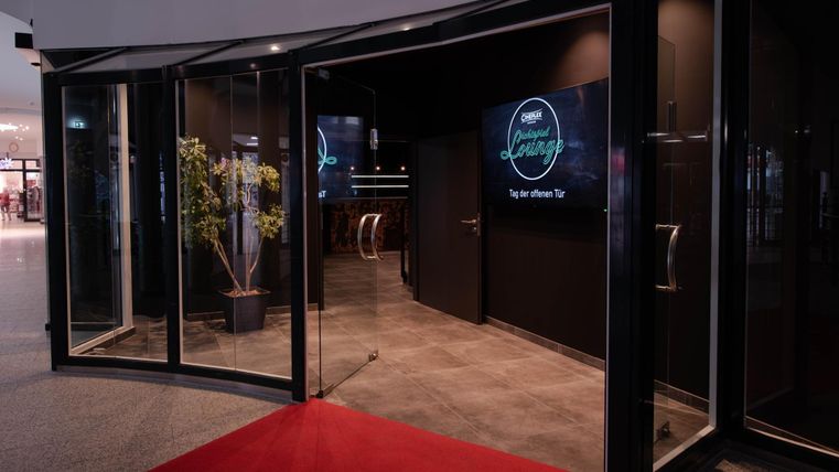 Een stijlvolle entree met glazen deuren en een rode loper. Binnen zijn moderne schermen en een gezellige sfeer te zien.