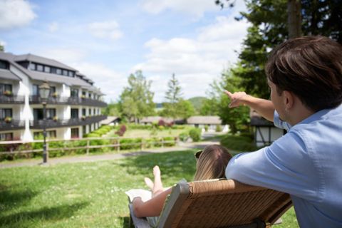 Ein Paar sitzt entspannt auf einer Liege und schaut auf eine schöne Landschaft. Im Hintergrund sind moderne Gebäude und viel Grün zu sehen.