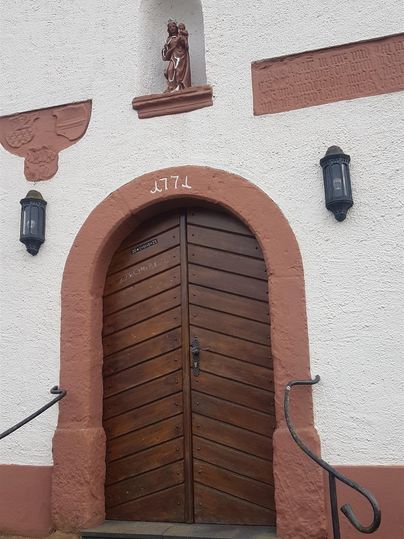 Eine rustikale Holztür mit geschwungenen Formen und einem hölzernen Figurenrelief darüber. Die Tür ist von einer weißen Wand und roten Akzenten umgeben.