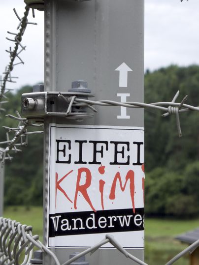 Zaun mit Stacheldraht und Schild 'Eifel Krimi Wanderweg' in ländlicher Umgebung.