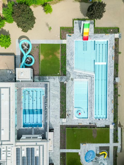 Ein Überblick über ein Schwimmbadgelände mit mehreren Becken und einer Riesenrutsche. Dabei sind grüne Rasenflächen und Spielgeräte sichtbar.