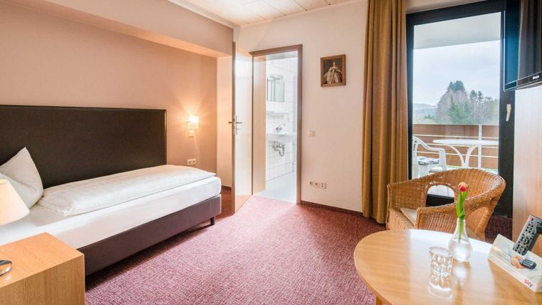 A bright hotel room with a single bed and a cozy armchair. There is a small table and access to a balcony.