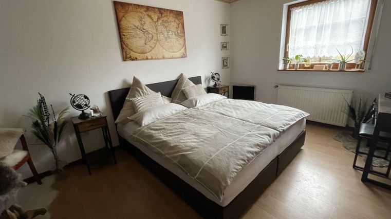 Een gezellige slaapkamer met een groot bed en een warme, uitnodigende sfeer. Aan de muren hangen decoratieve schilderijen, en het licht komt binnen via een raam met gordijnen.