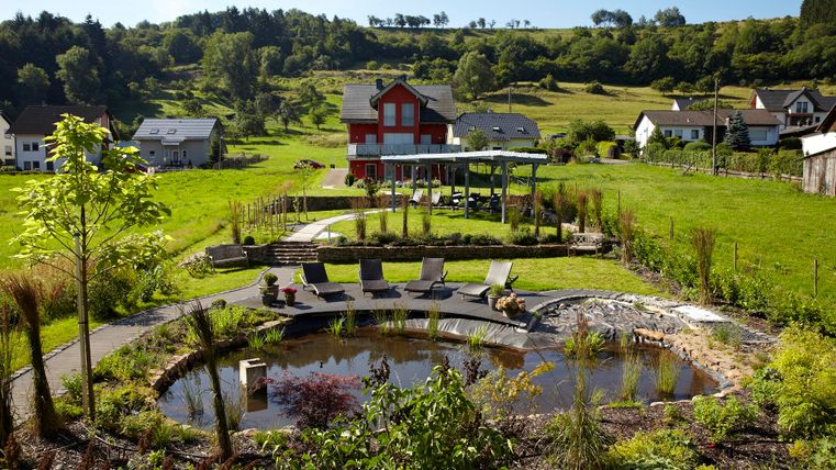 A picturesque garden with a round pond and deck chairs. In the background, green hills and a red house are visible.