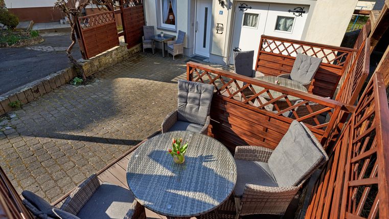 A cozy outdoor area with a round table and comfortable chairs. Surrounded by wood paneling and a beautiful plant pot.