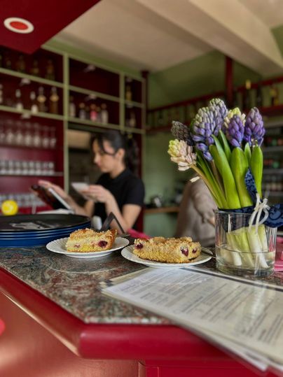 Zwei Teller mit Kuchen stehen auf einem Tisch in einem Café. Im Hintergrund ist eine Mitarbeiterin zu sehen, die eine Speisekarte liest, während eine Vase mit Hyazinthen auf dem Tisch steht.