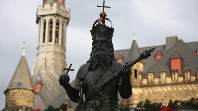 Eine Statue eines König mit Krone und Kreuz in der Hand. Im Hintergrund sieht man beeindruckende mittelalterliche Gebäude.