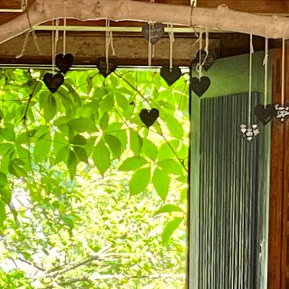 Ein Fenster mit hängenden Herzen aus Holz und grünen Blättern im Hintergrund. Licht strömt durch das Fenster und schafft eine gemütliche Atmosphäre.