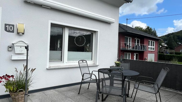 Eine Terrasse mit einem Tisch und Stühlen. Im Hintergrund sind weitere Gebäude und ein bewölkter Himmel zu sehen.
