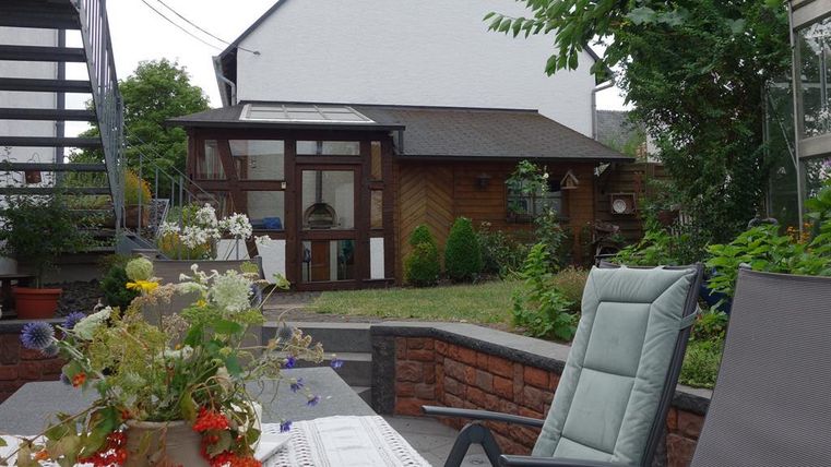 Een gezellige tuin met een tafel en stoelen. Op de achtergrond zie je een houten gebouw en weelderige planten.